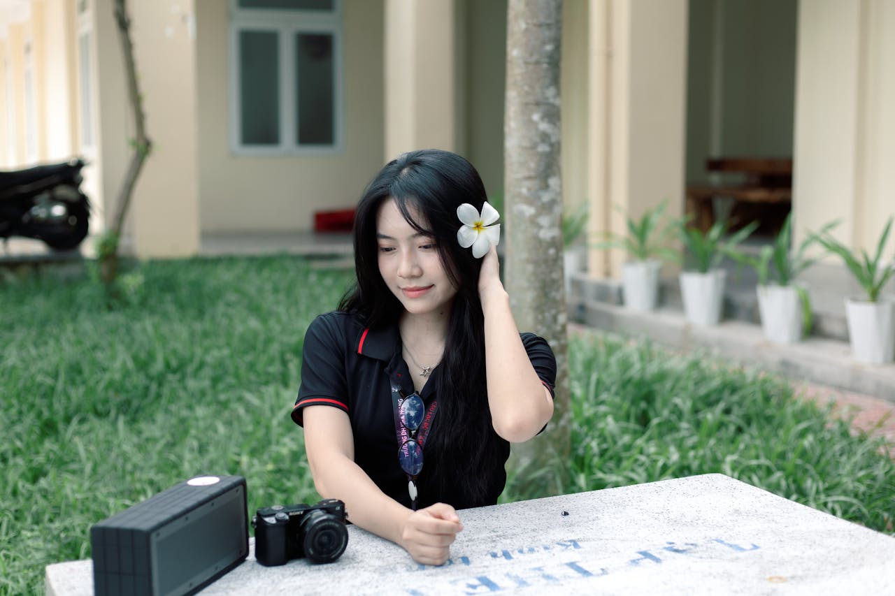 A young woman with a flower in her hair relaxes outdoors with a camera and speaker.